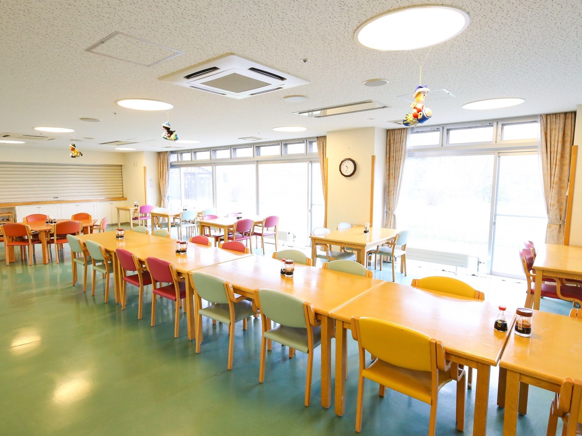 食堂に多くのテーブルと椅子が置かれている。椅子の座面や背もたれは、いろいろな色がある。大きな窓からの採光で明るい。