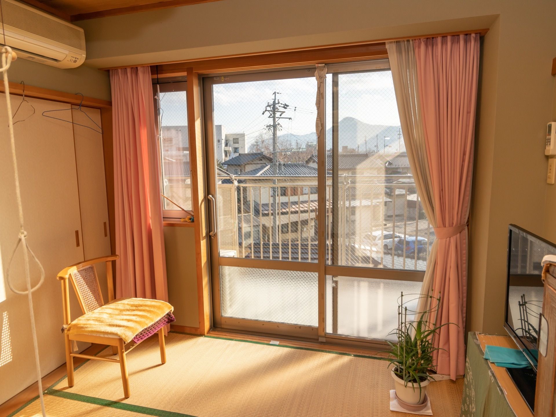 畳の部屋に、椅子とテレビが置かれている。ピンクのカーテンのかかった窓からは日光が良く入っている。エアコンが設置されている。