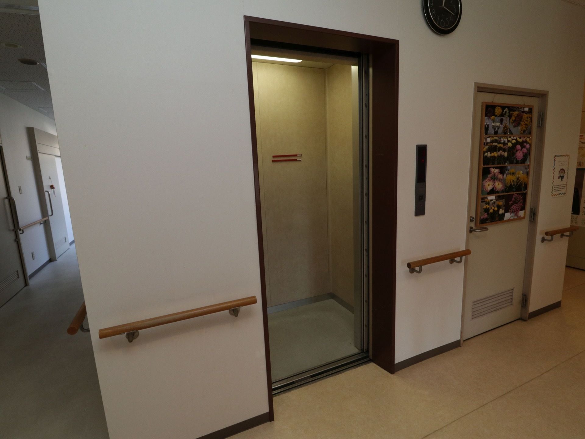 廊下にエレベーターが１基設置されている。周りの壁には手すりが設置されており、時計が取りつけられている。