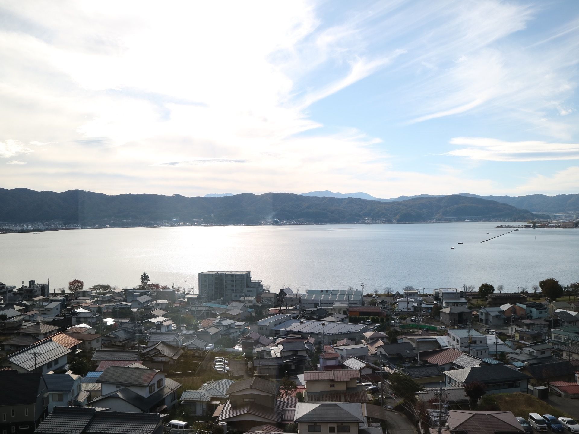 晴天の日に施設から見た景色である。住宅地が眼下に広がり、先には湖が開け、山々が奥に連なっているのが見える。