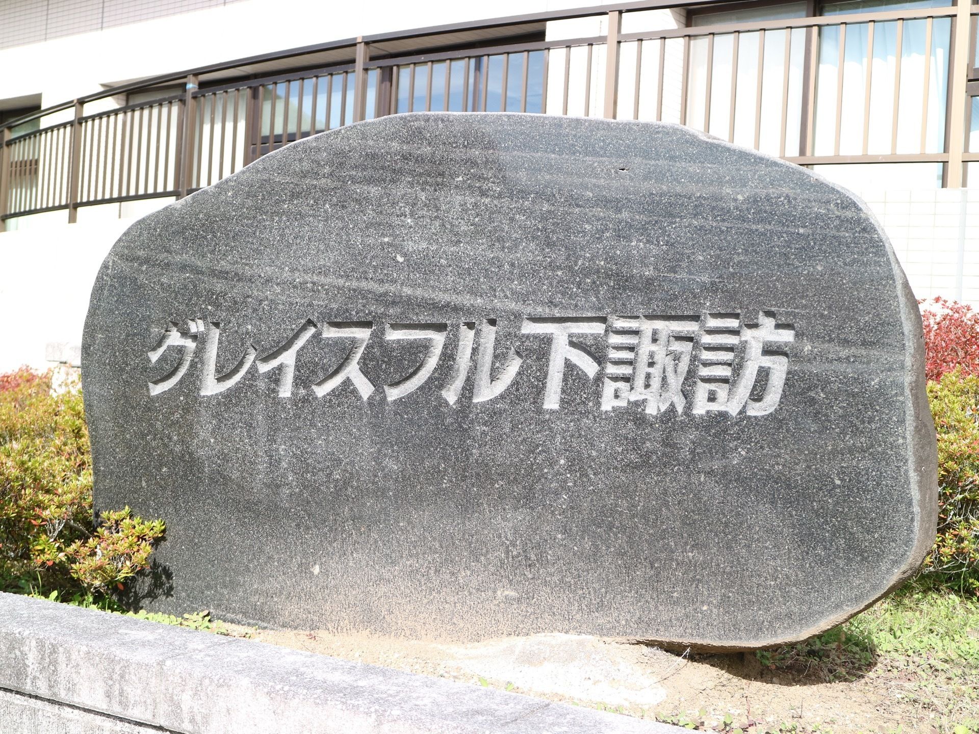 ダーク色の大きな1枚岩の断面に、施設名称が横一列に彫り込まれ、施設建物前の植え込みに設置されている。