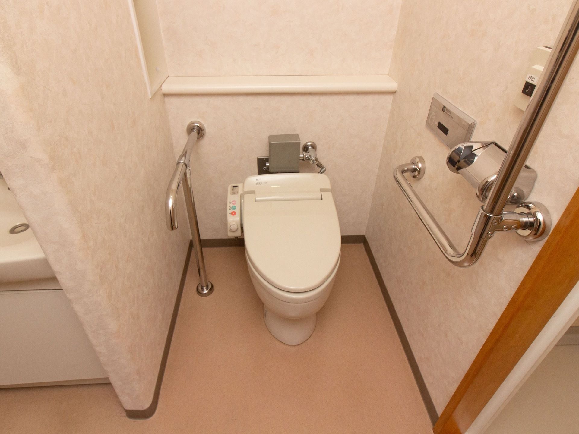 トイレの背面には物が置ける棚があり、小物を片しておくことができる。壁の両側には手すりがついて、緊急用のボタンも取りつけられている。