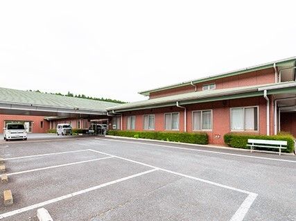 白線が引かれた広い駐車場が付いたエンジ系の外壁の建物。建物の周りには低木が植えられ、ベンチも置かれている。