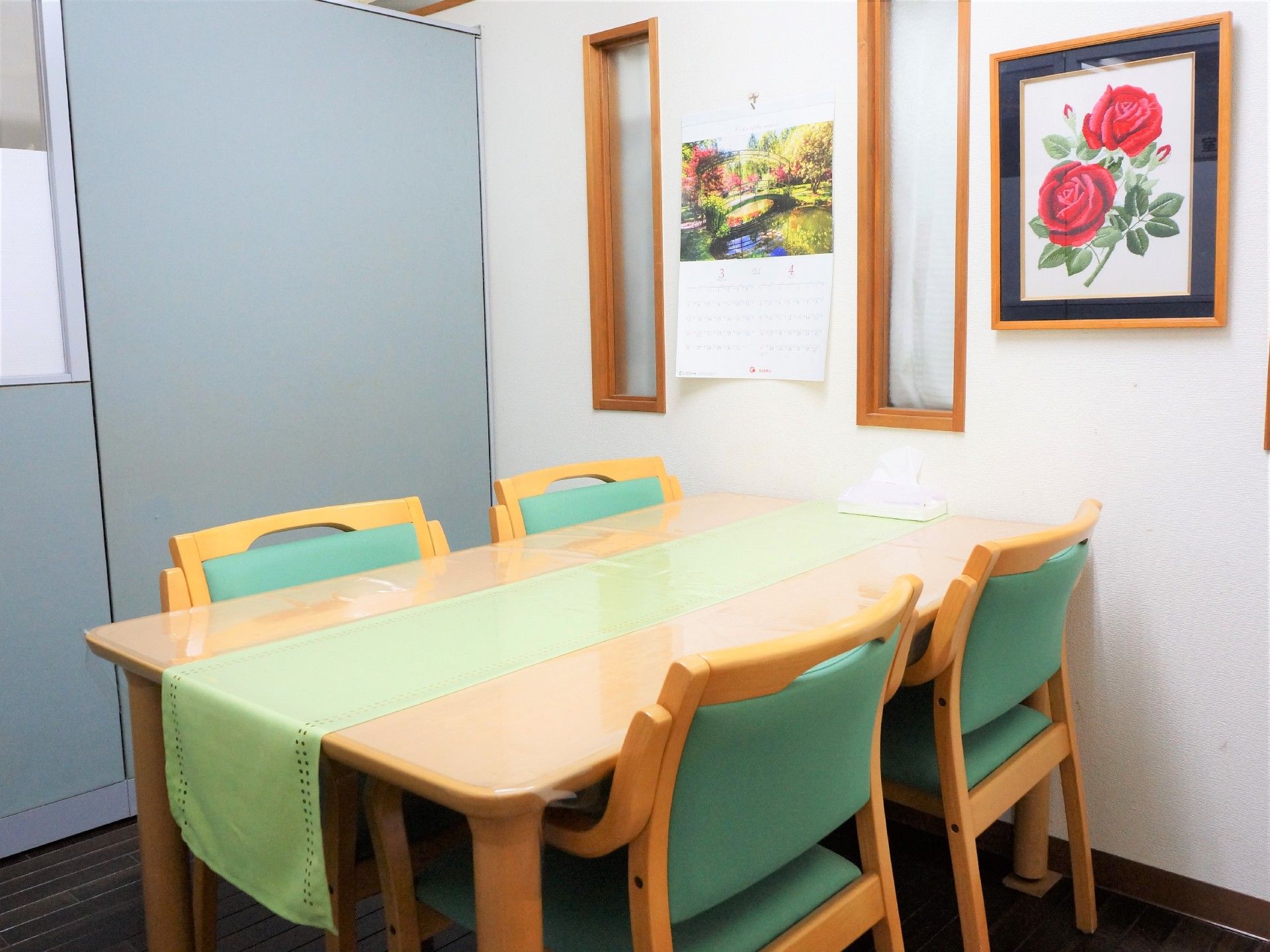 部屋には4つの椅子と大きなテーブルが設置されており、細長いテーブルクロスが置かれている。カレンダーや絵画が壁にかけられている。
