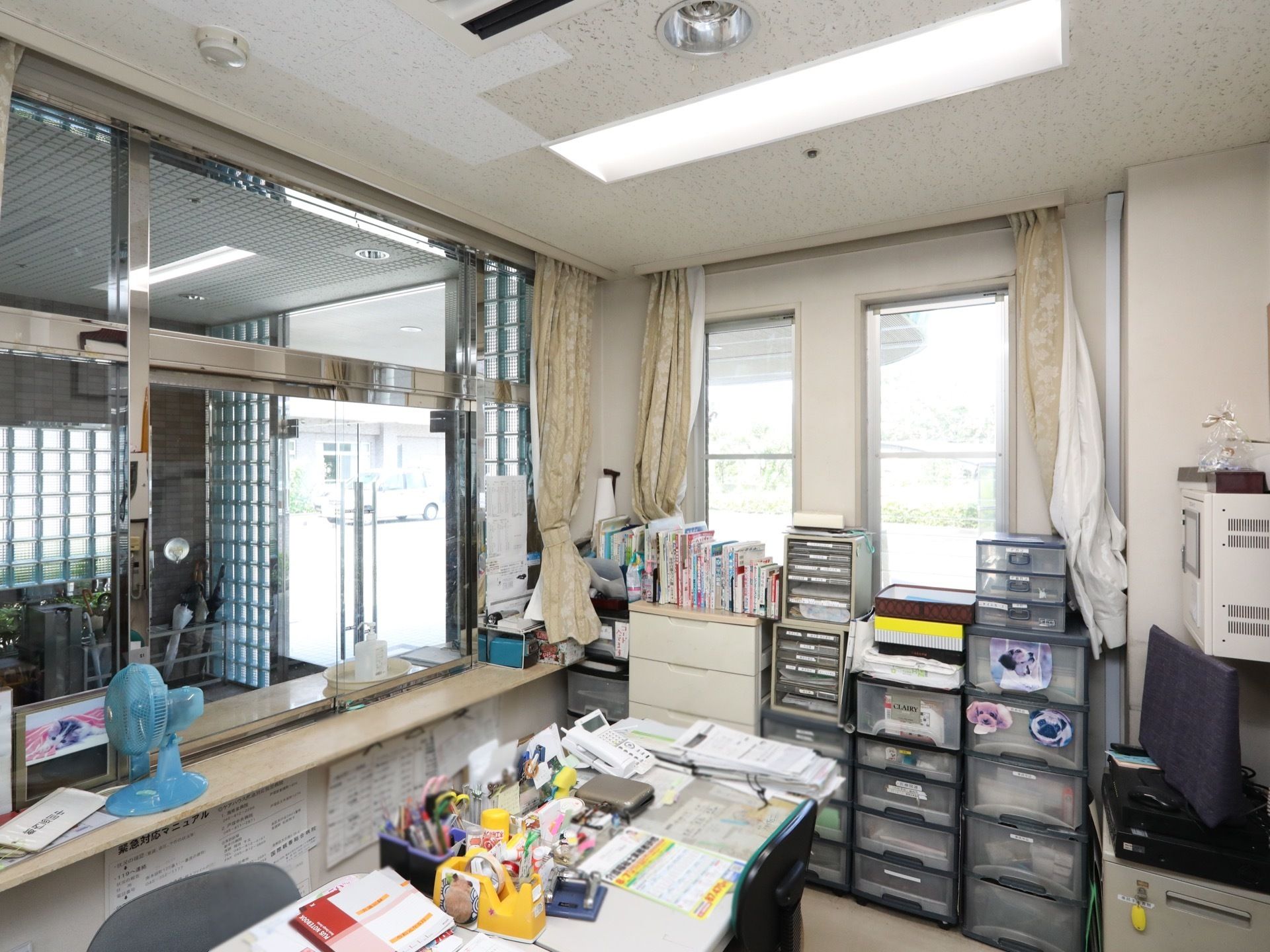 部屋の中央には机が置かれており、椅子が用意されている。その上には資料や文房具が見えている。電話機がある。