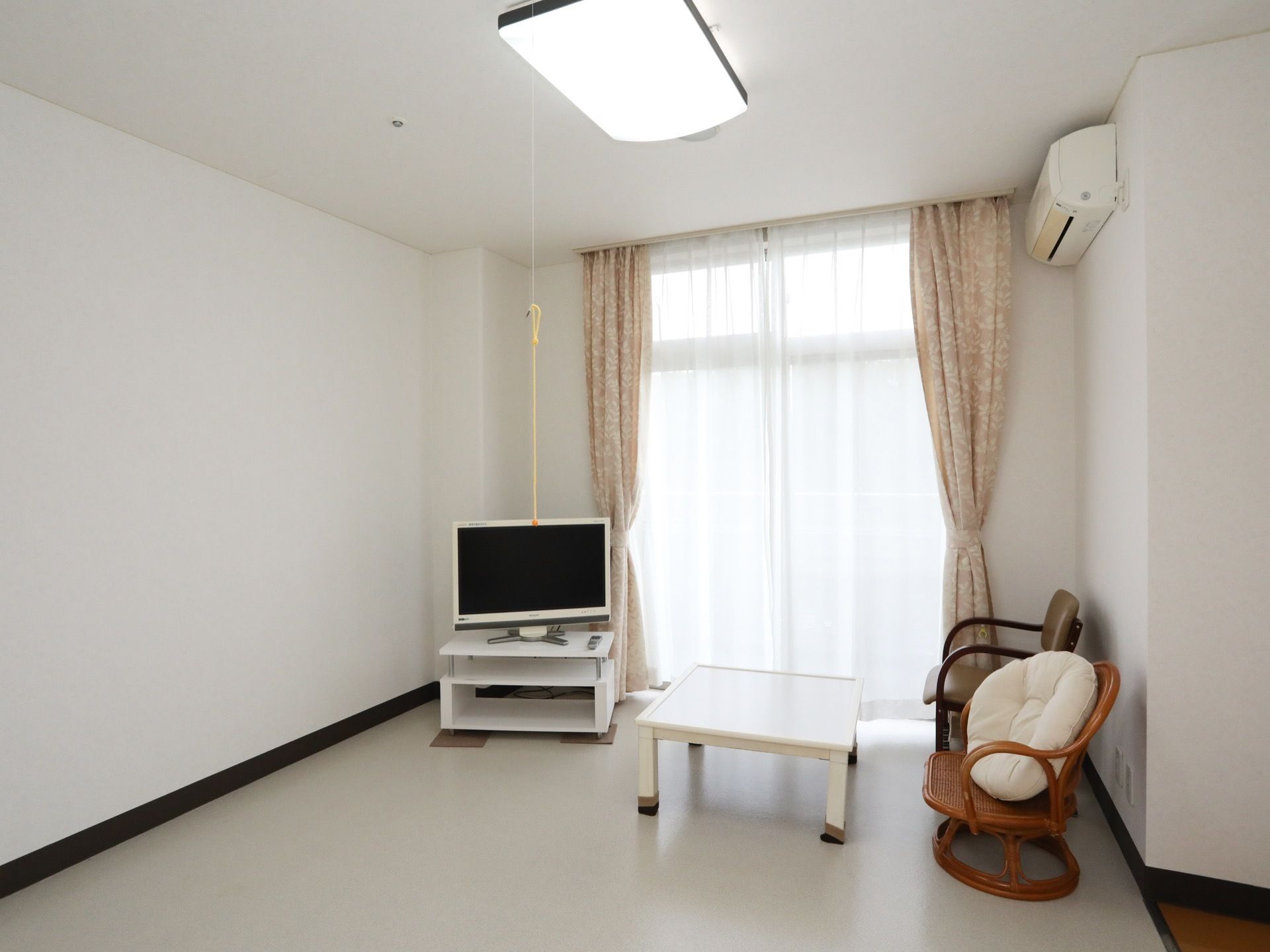 大きな窓のある明るい居室で、床はフローリングとなっている。テレビや椅子、テーブルなどの家具が配置されている。