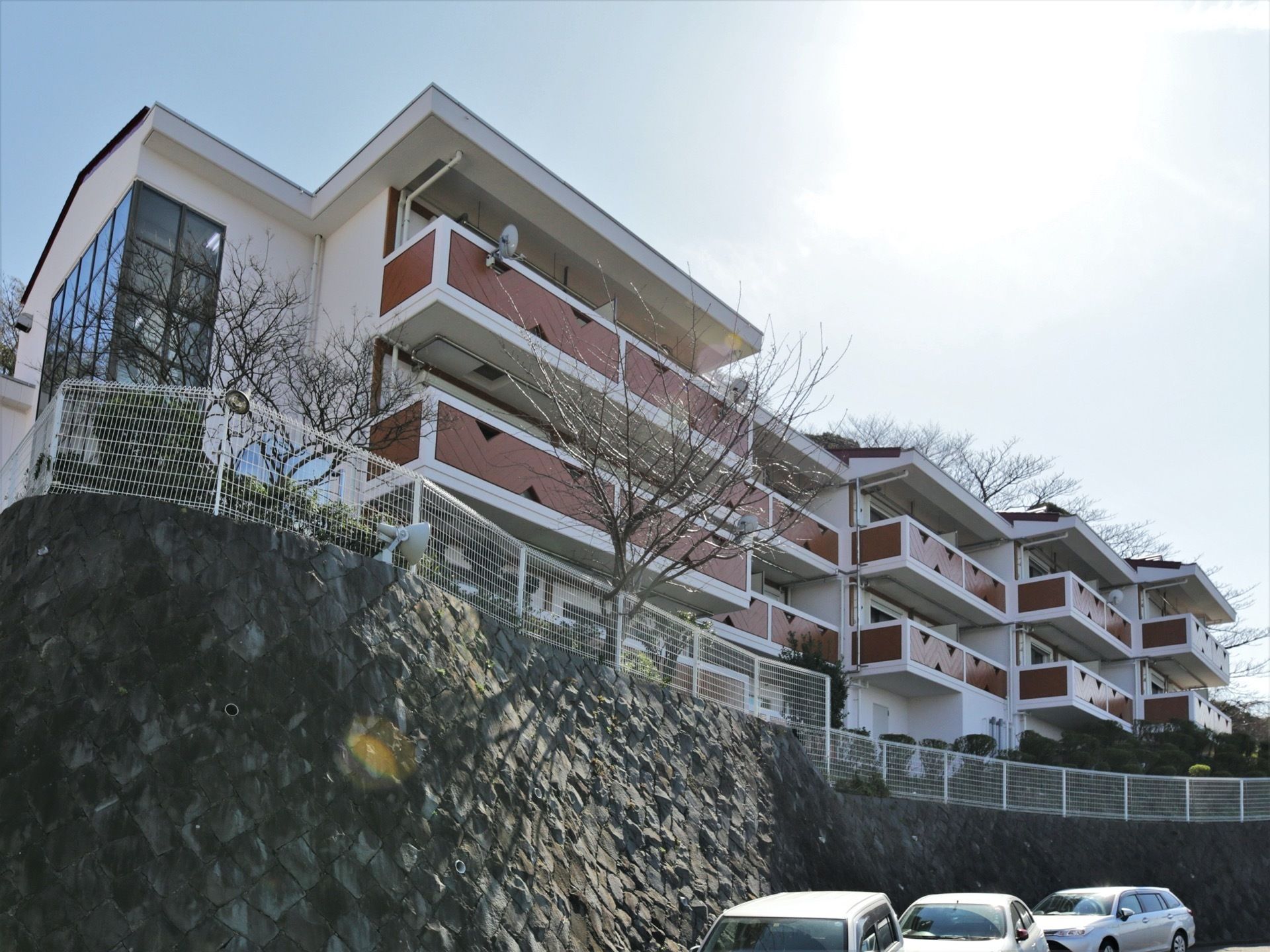 石組み壁の小高い地に立地している３階建の施設である。各居室にベランダが設けられている。眼下の駐車スペースには、乗用車が停車している。