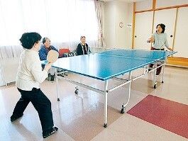 卓球を楽しんでいる女性たちの様子。レースカーテンとカーテンが付いた大きな窓のそばには２名観戦をしている人もいる。