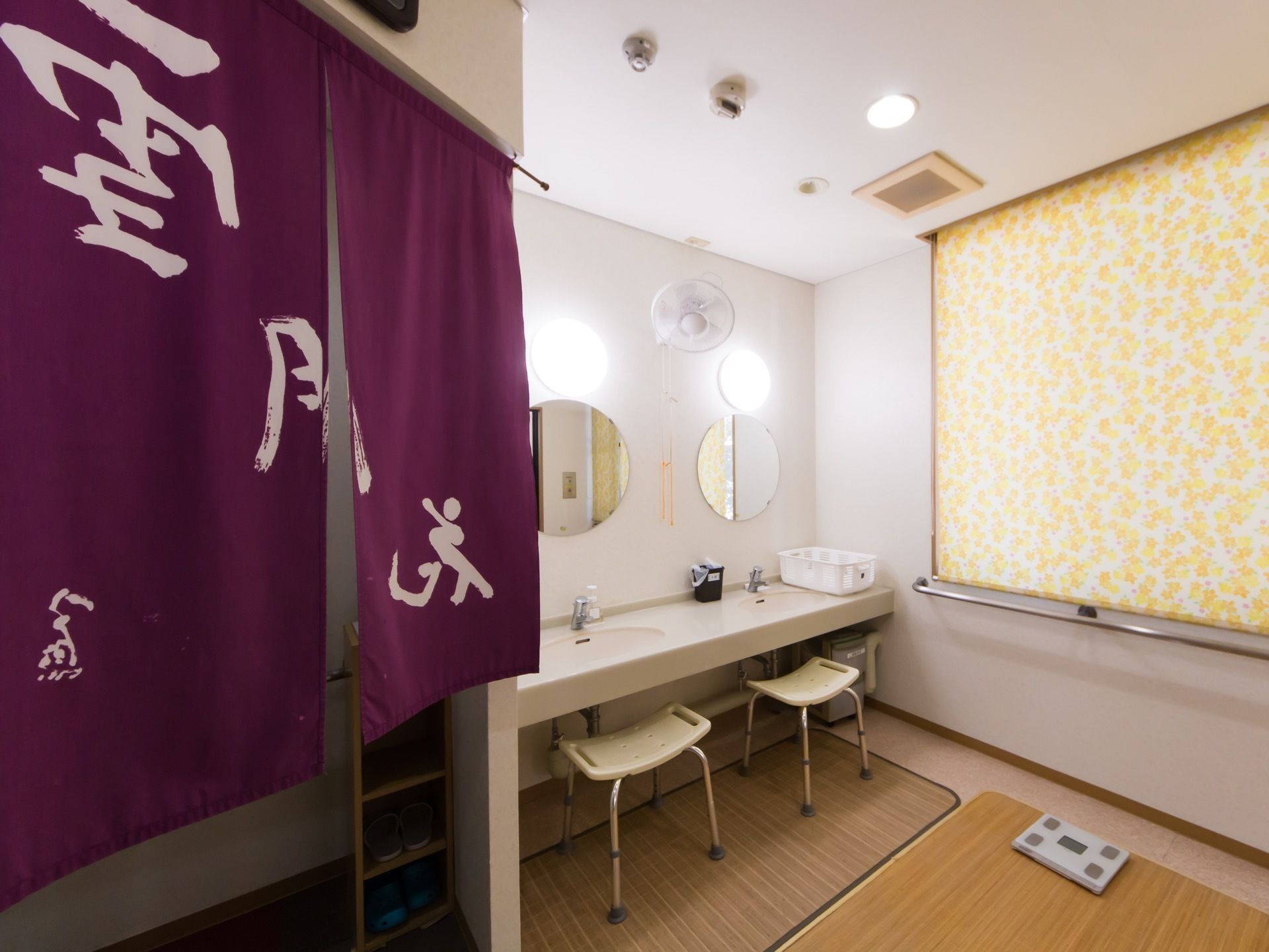 脱衣室の入り口に文字が入ったのれんが掛かっている。2人が使用できる洗面台や体重計が見える。明るい花柄のロールスクリーンがある。