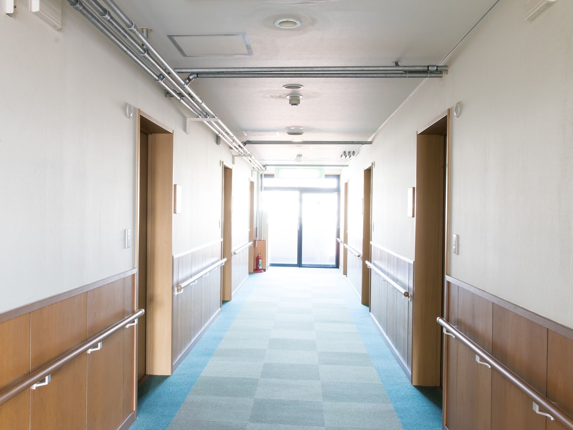 居室前の廊下は白と木調で温かみがある。床は滑りにくいカーペット張り。天井にスプリンクラー、廊下の突き当りに消火器が見える。