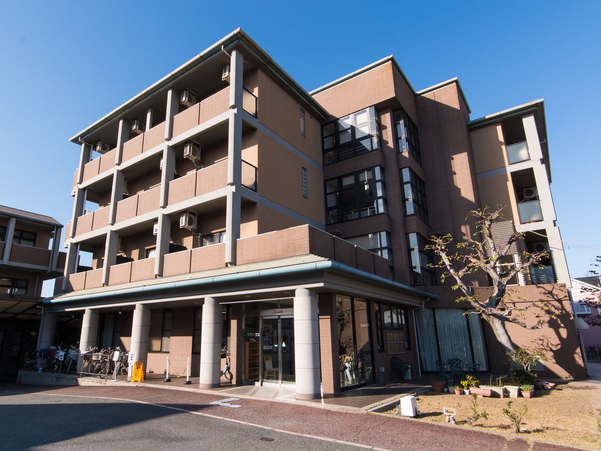 4階建ての大きな建物は重厚感がある。奥に駐輪場、各室外にエアコンの室外機が並んでいるのが見える。芝生の庭や木々が植えてある。