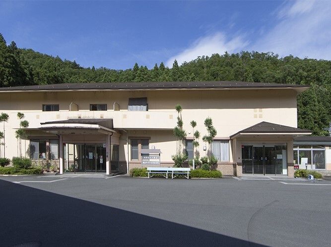 緑に囲まれた施設は広い駐車場が付き、2階建ての建物はベージュ系の外壁で、玄関が2カ所にある。