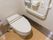 Ｌ型手すりや呼び出しボタンを完備したトイレである。使い捨ての便座シートホルダーが壁に設置されている。