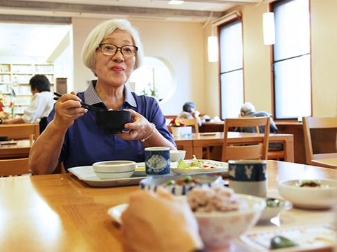 眼鏡をかけた高齢女性がお椀を持って微笑んでいる。女性の前のテーブルにはトレーに並んだ食事がある。