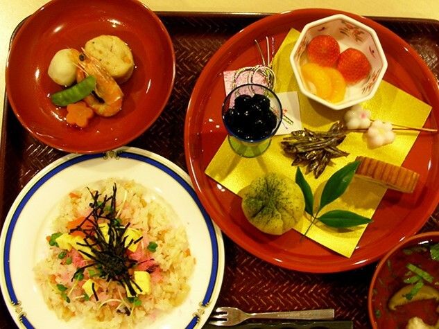 ちらし寿司や吸い物、煮物、多作りや黒豆などの正月料理がちょっとずつ盛られた皿がある。