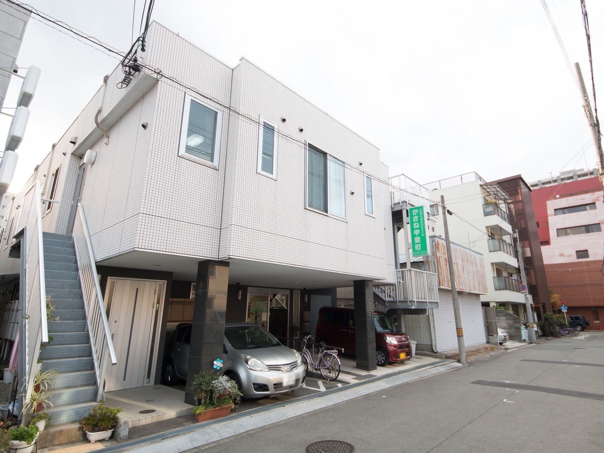 白い建物の外壁には緑の看板が設置され、1階部分の駐車場には車が止められている。