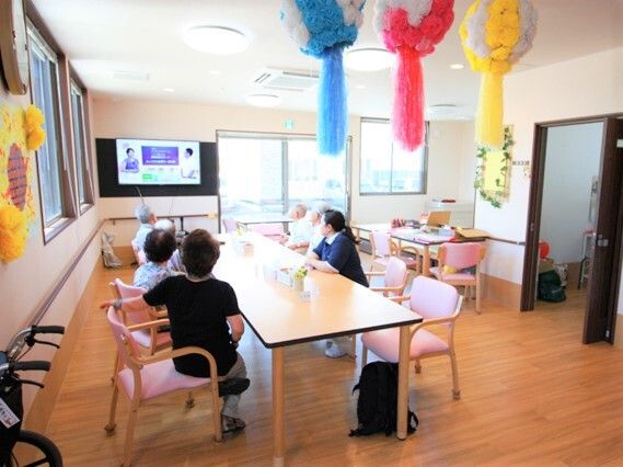 天井には夏の七夕飾りが飾られ、壁にはヒマワリの壁画が飾られている。食堂のダイニングテーブルに数名のスタッフと入居者が座り、テレビを見ている。
