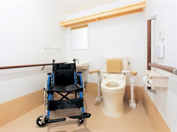 車いすが置かれた広いトイレ。手すりや背もたれが付いたトイレが右側にあり、車いすの後ろには手洗い場がある。
