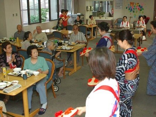 手作りの花が飾られた傘を持って踊っている女性たちの踊りを熱心に見ている高齢者たち。