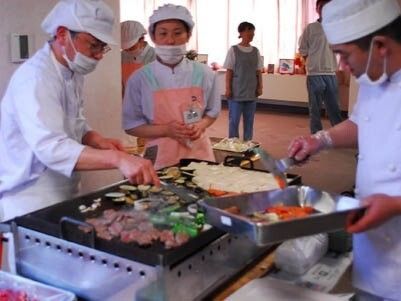 厨房用の白衣を着た男性スタッフ２名とエプロンを付けた女性スタッフが鉄板焼きを作っている。