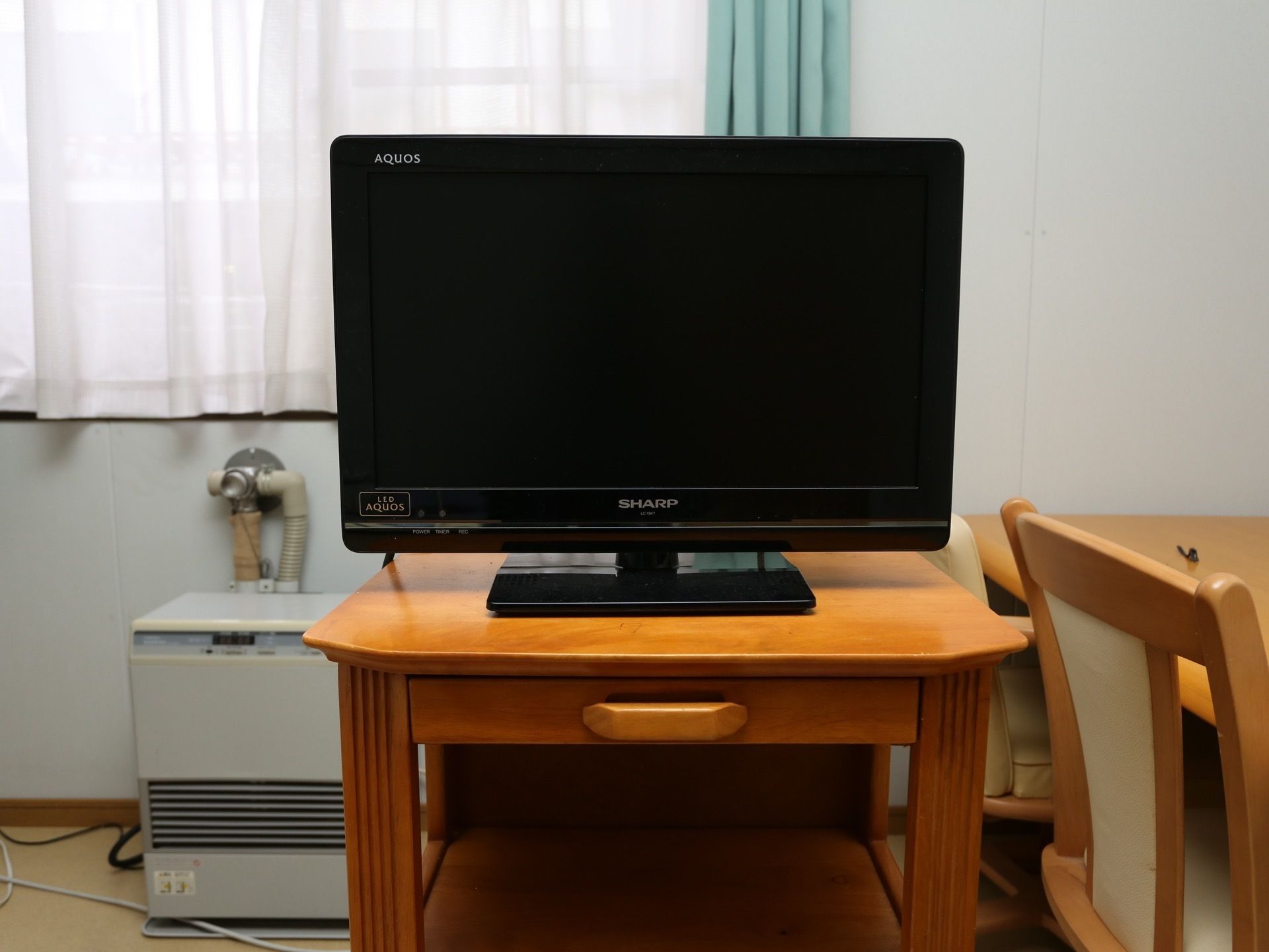 木製の台の上に黒いテレビが置かれている。後ろには窓があり、白いレースとグリーンのカーテンがかけられている。
