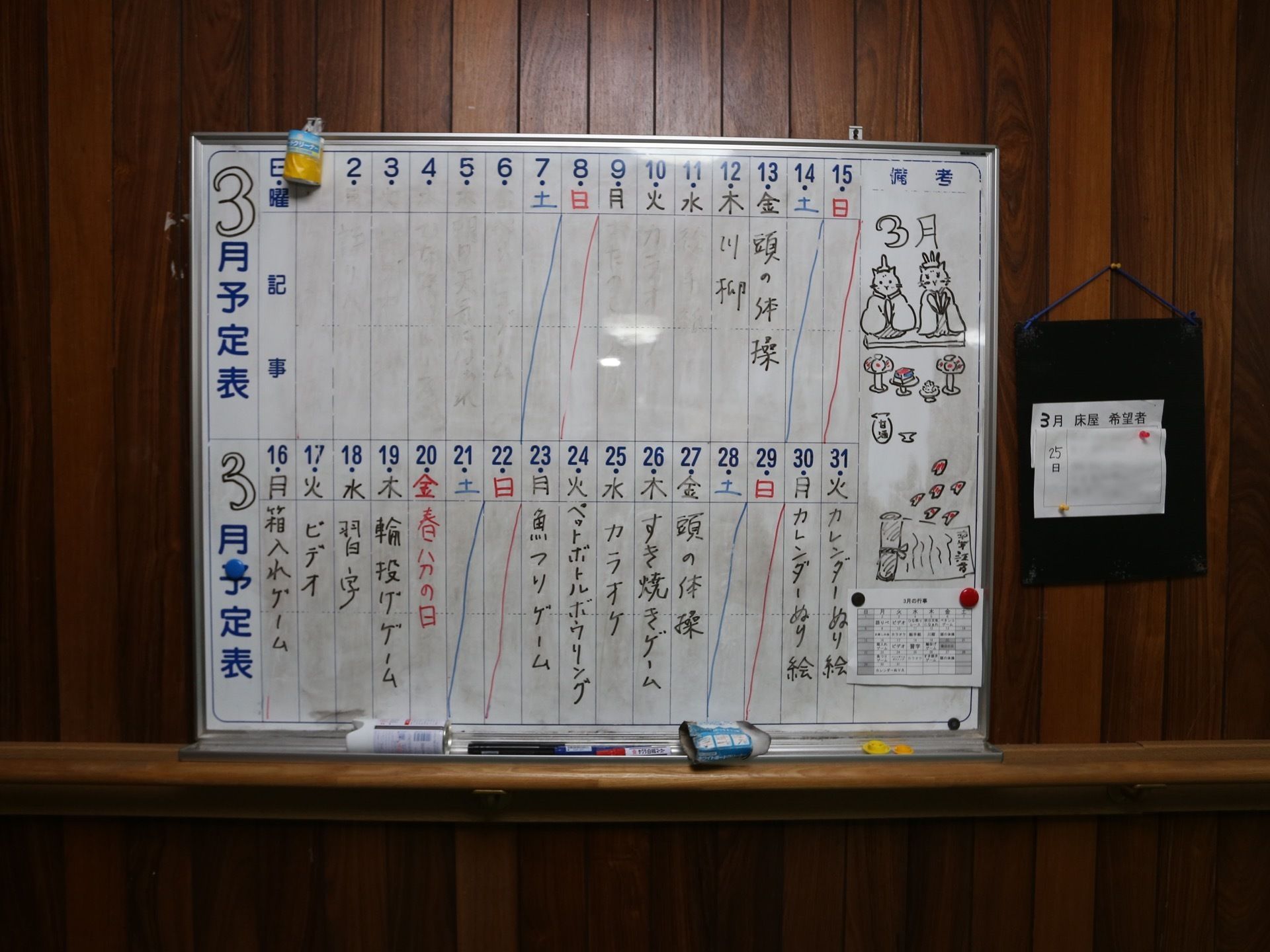 濃い色合いの木目調の壁にホワイトボードが付けられている。ボードには、３月の予定が書き込まれている。