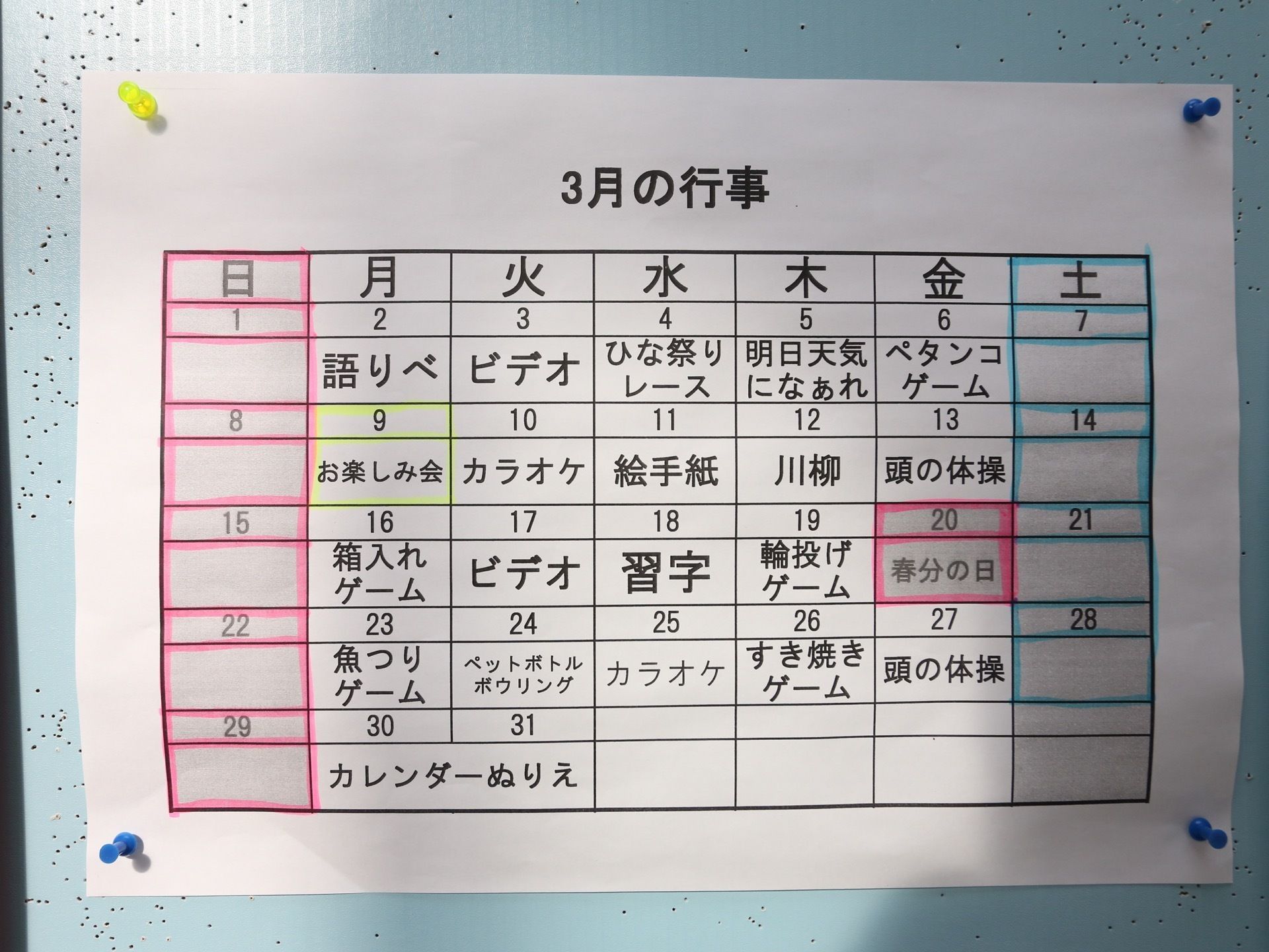 壁に３月の行事がプリントされた紙が貼られている。日付ごとにイベントが記載され、日曜と祝日はピンク、土曜は青いペンで囲んである。