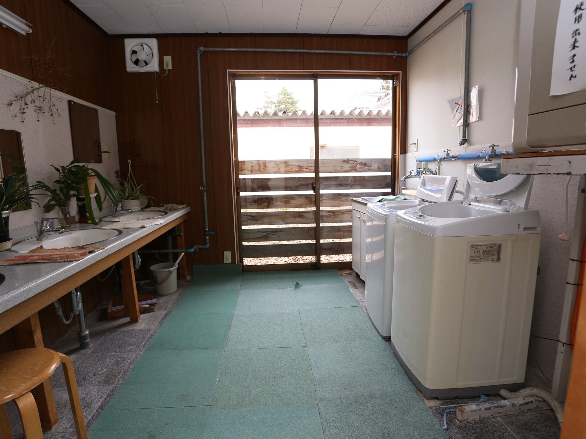 白い縦型洗濯機が２台壁際に並んでおり、向かい側に洗面カウンターが設置されている。突き当たりに掃き出し窓があり、床にマットが敷かれている。
