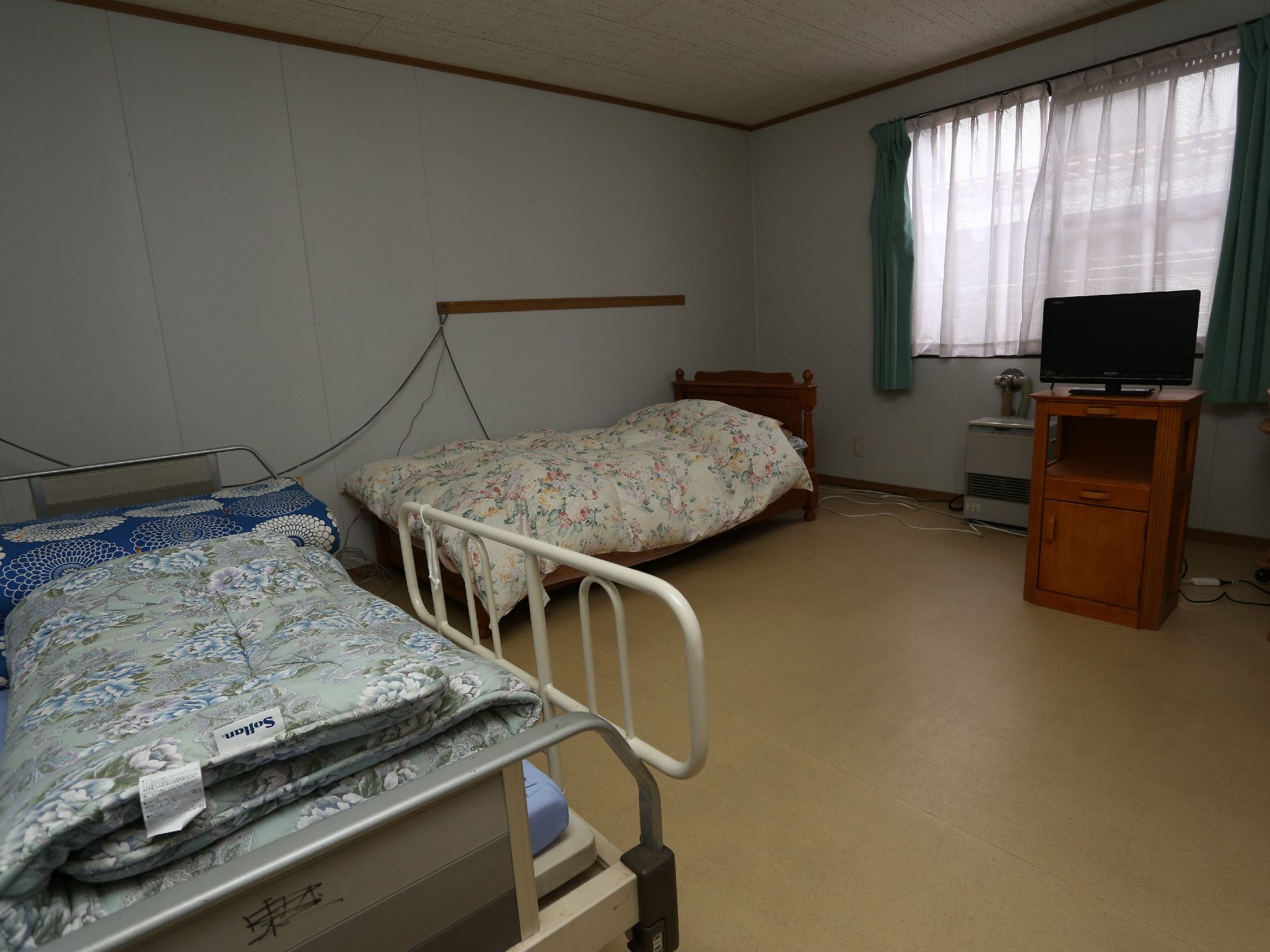 右側に窓があり、その左に花柄の布団のベッド、さらに左手前に青い花柄の布団のベッドがある居室