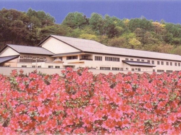 沢山のピンクのつつじの花に囲まれた三角屋根が付いた白い建物。その奥には山が連なっている。