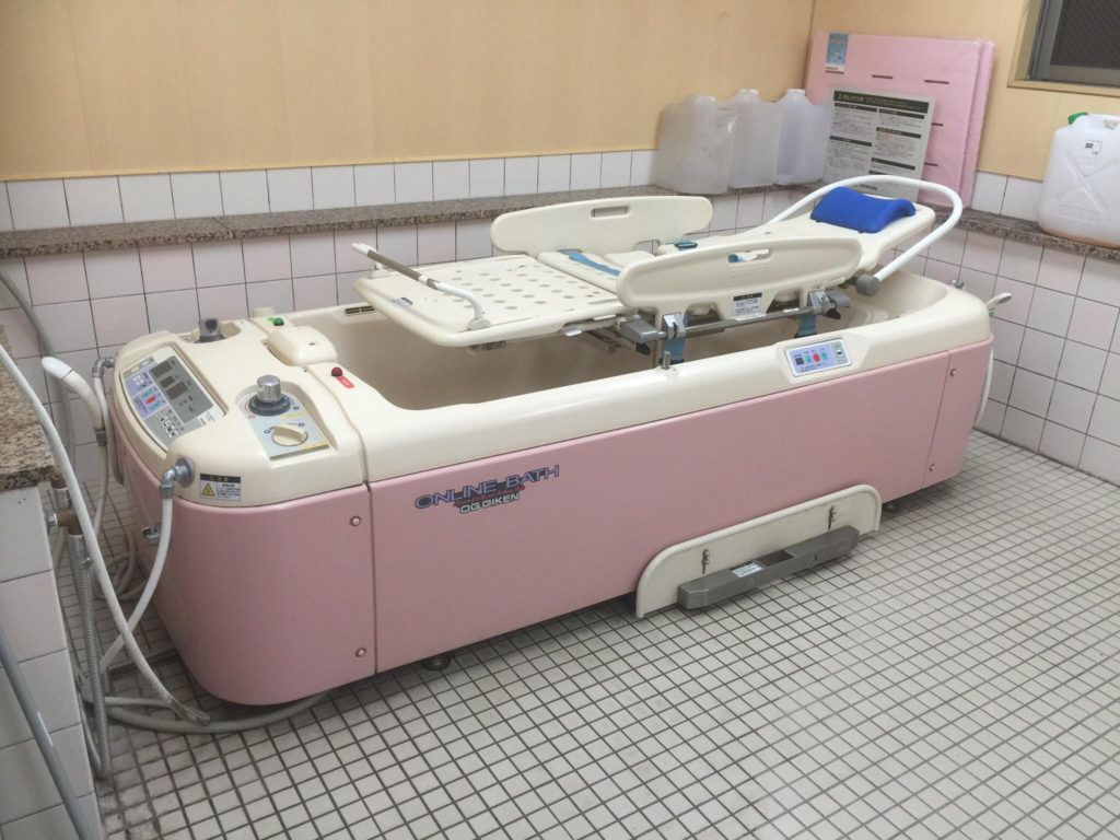 ピンクの寝台浴。寝たまま入浴できる設備。浴室は白いタイルばりで棚には大きなボトルなどが置かれている。