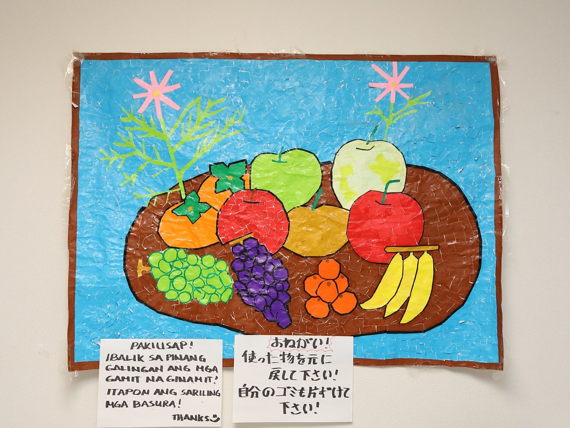 さまざまなフルーツと花を描いたちぎり絵。鮮やかな色紙で表現されており、存在感のある作品である。下には注意書きが張り付けられている。