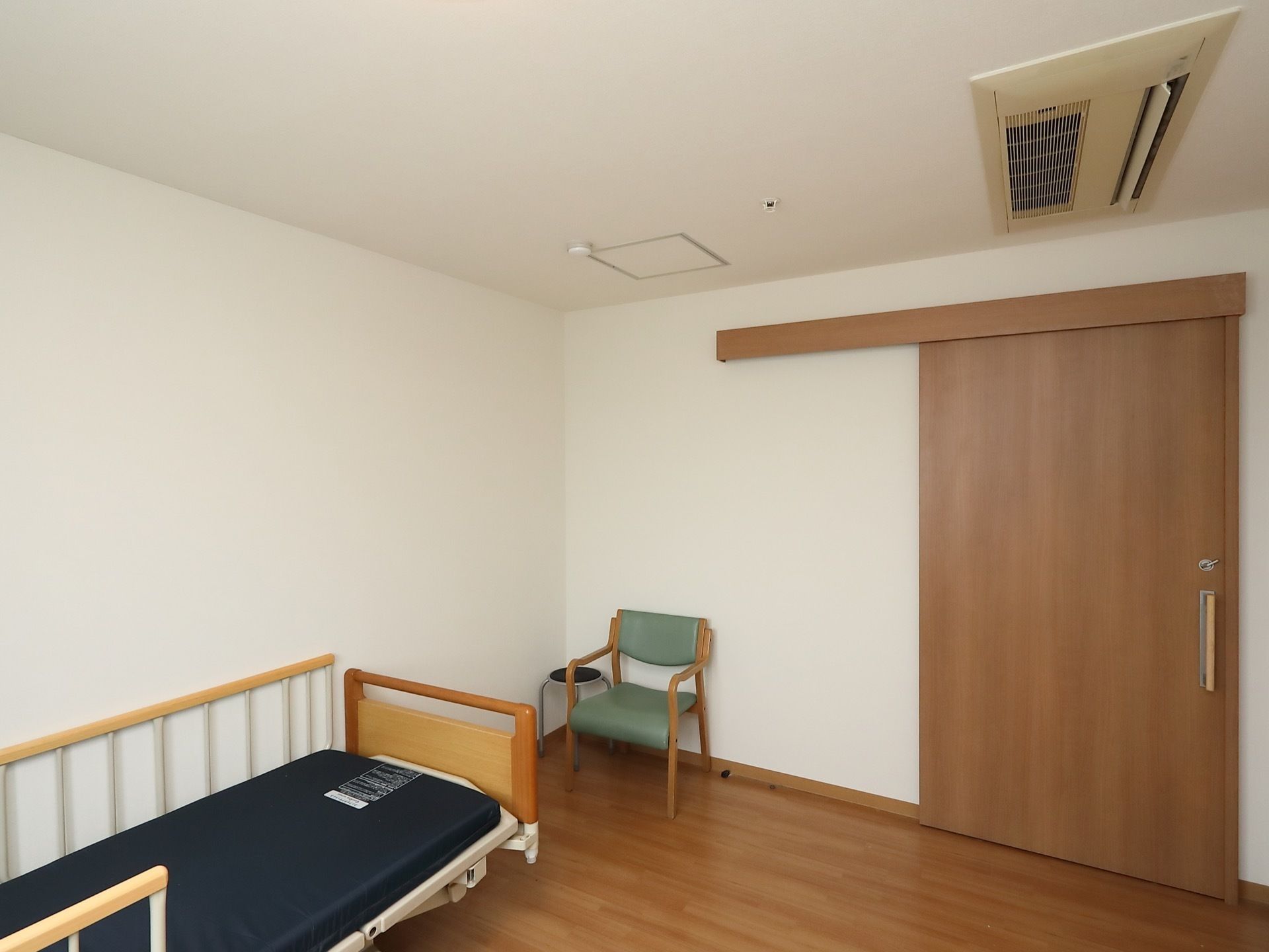 居室の入り口は引き戸である。天井にはエアコンのほか火災報知器が埋め込まれている。介護用ベッドや椅子、建具は木調でまとめられている。