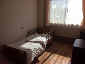 レースカーテンとカーテンが付いた窓がある部屋には寝具付きのベッド、緊急通報装置が付いている。