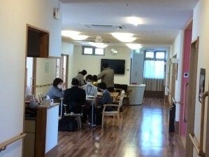 フローリングのアットホームな雰囲気のリビングダイニングでスタッフや入居者が集まって何かをしている。
