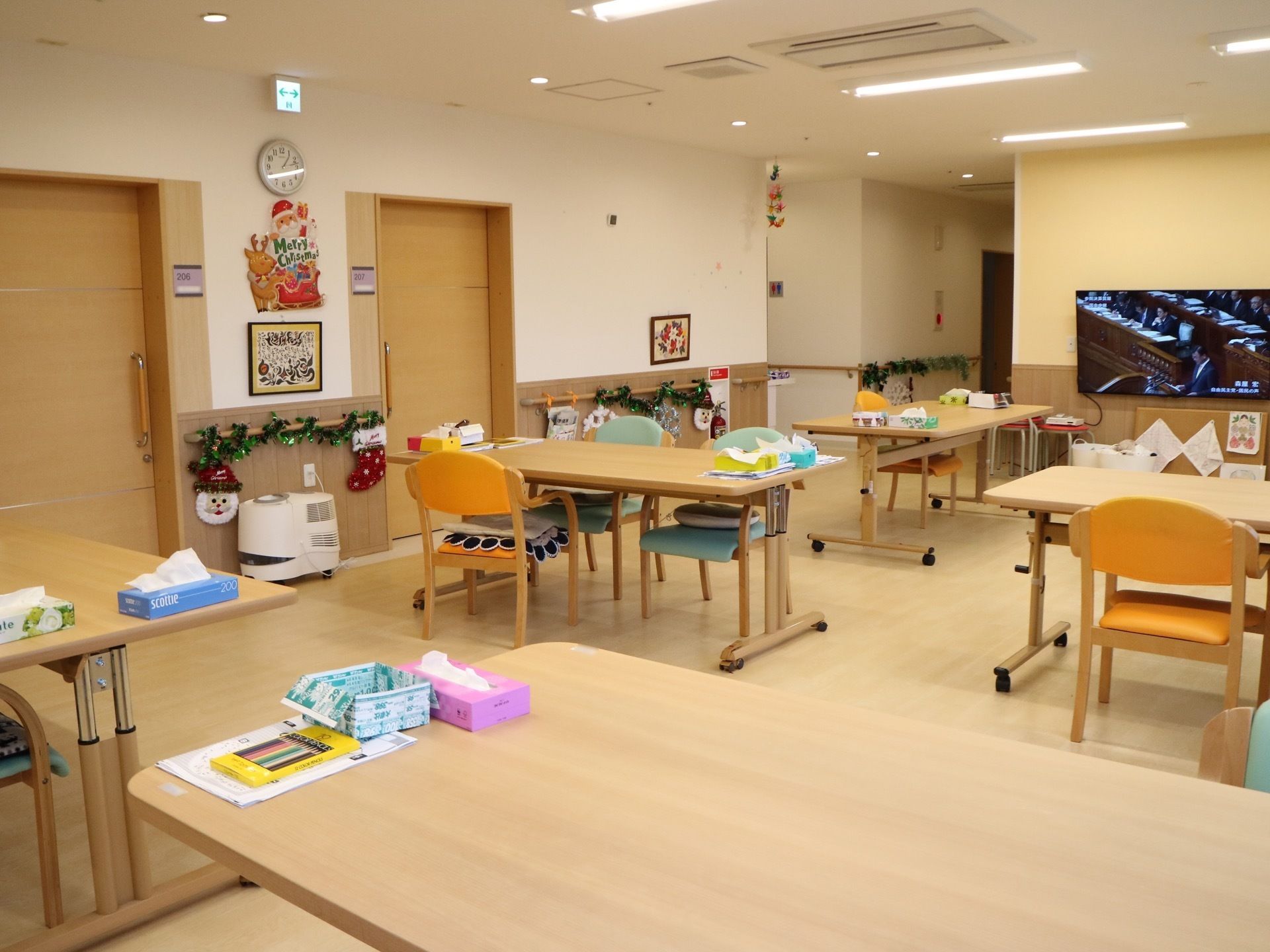 ダイニングテーブルが並び、その上には色鉛筆やティッシュが用意されている。壁面にはクリスマスの飾りが付けられ、テレビが設置されている。
