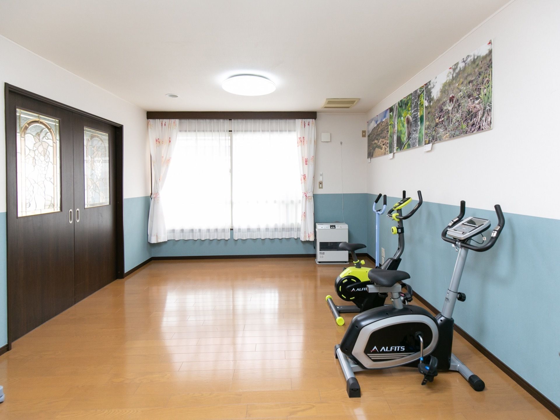 館内にある機能訓練室の画像である。右手の壁際にエアロバイクが２台置かれている。壁に風景画が貼られている。