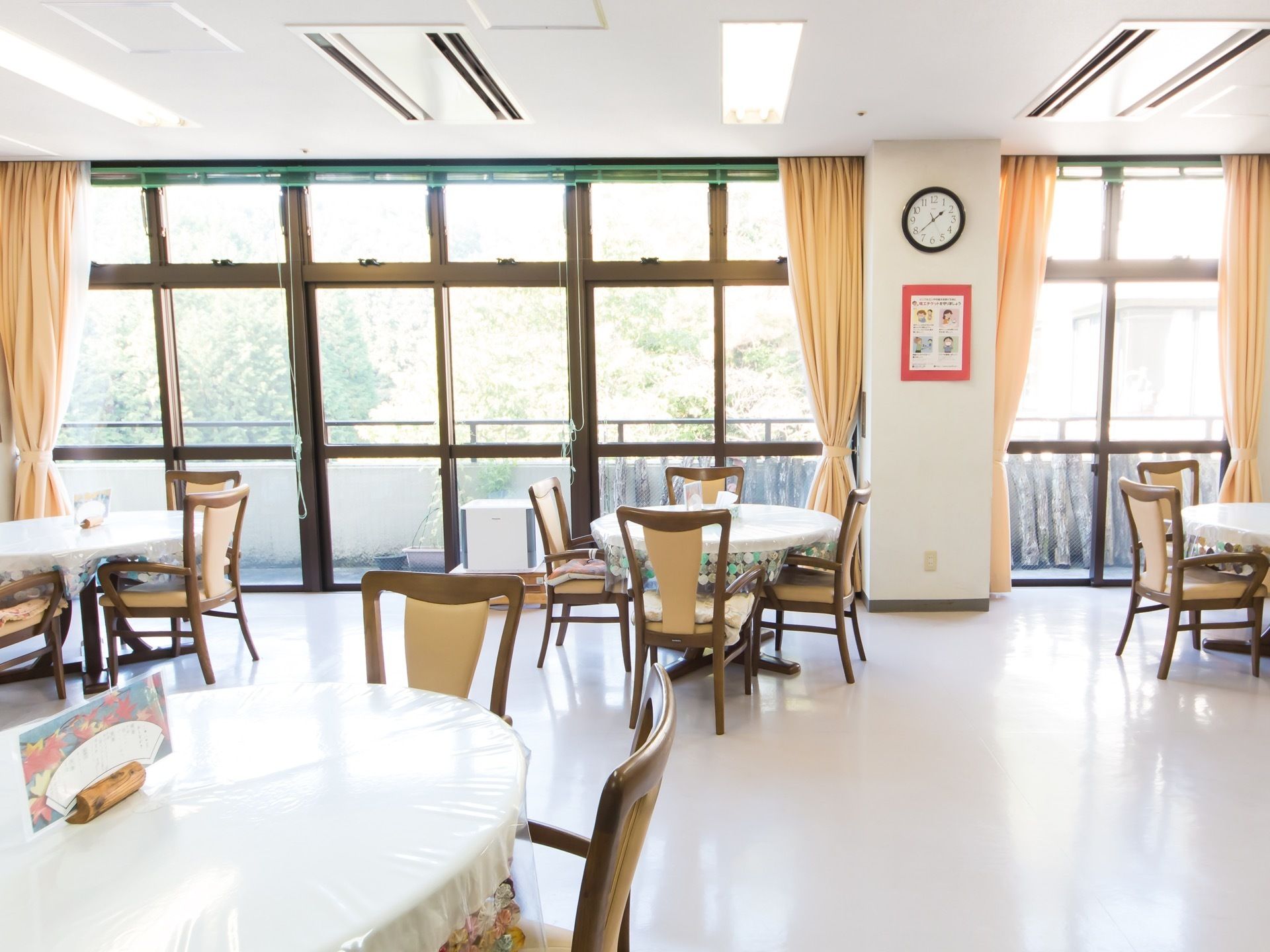 食堂に、丸テーブルがゆったりと間隔を空けて置かれている。その周りに、ひじ掛けと大きな背もたれのついた椅子がある。