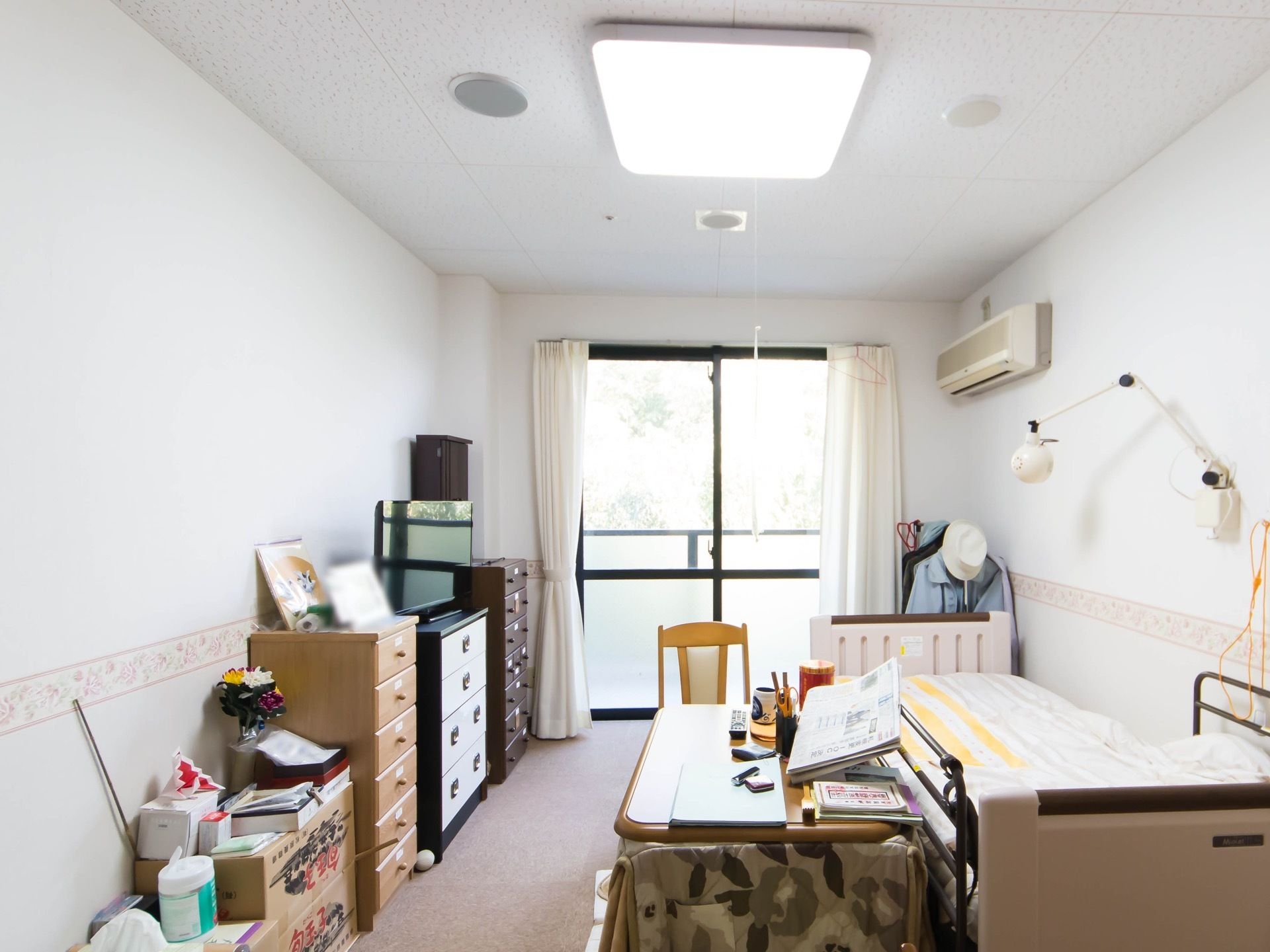 居室の壁際に、収納棚が３つ並べて置かれている。その真ん中の棚の上にテレビが載っている。天井の照明から紐がぶら下がっている。