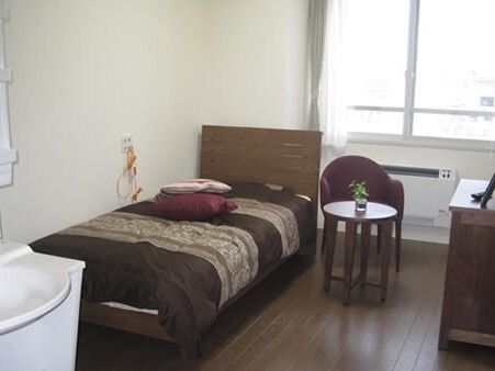 ブラウン系の寝具がセットされた一人用のベッドのそばには緊急通報装置があり、窓のそばにはミニテーブルセットがある。