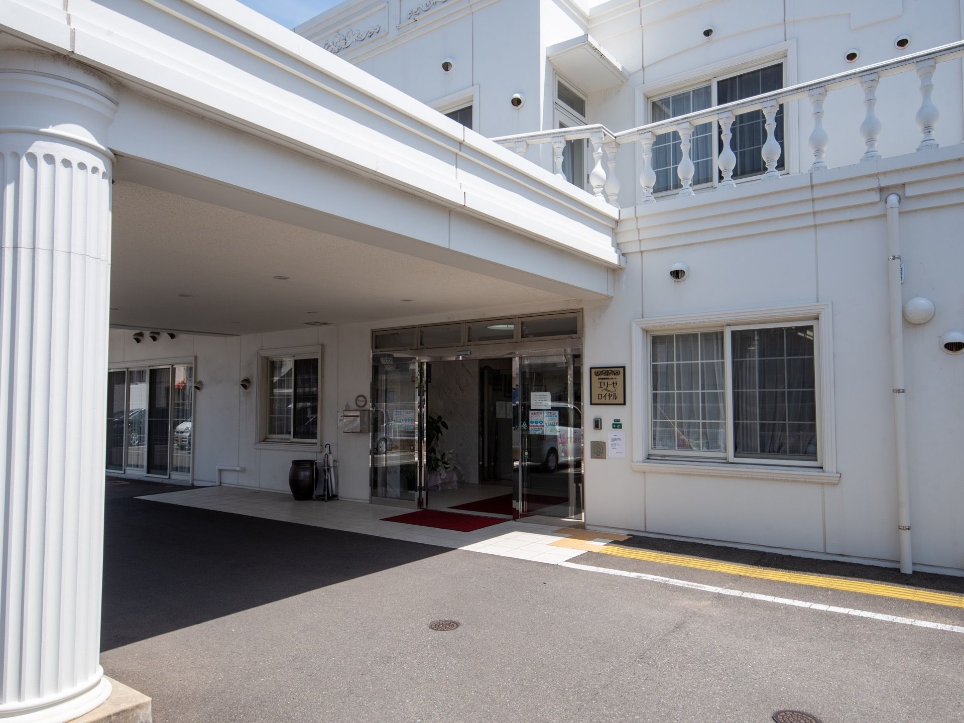 白い壁の２階建ての施設の玄関がある。玄関先は広いルーフで覆われている。地面は段差がなく、舗装されている。
