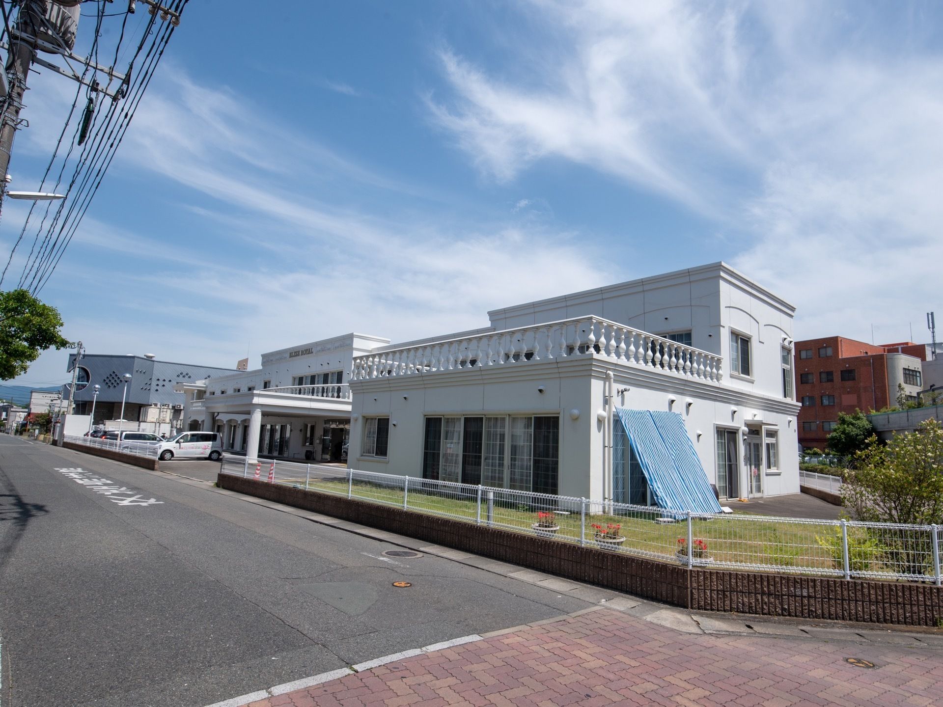 2階建ての白い大きな建物がある。2階部分は一部テラスになっている。敷地内には芝生の庭があり、背の低いフェンスで囲まれている。