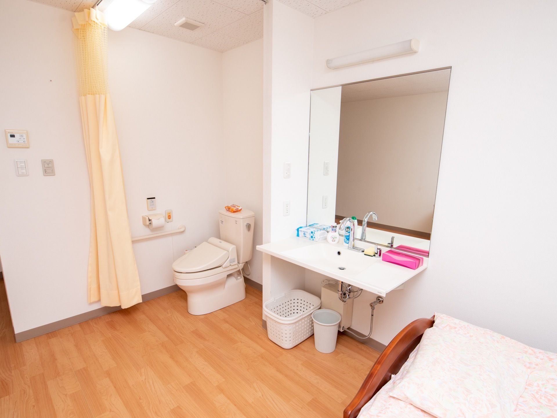 居室に備わっている洗面台。すぐ隣には、カーテンで仕切られたトイレがある。洗面台の正面には大きな鏡があり、照明器具が付いている。