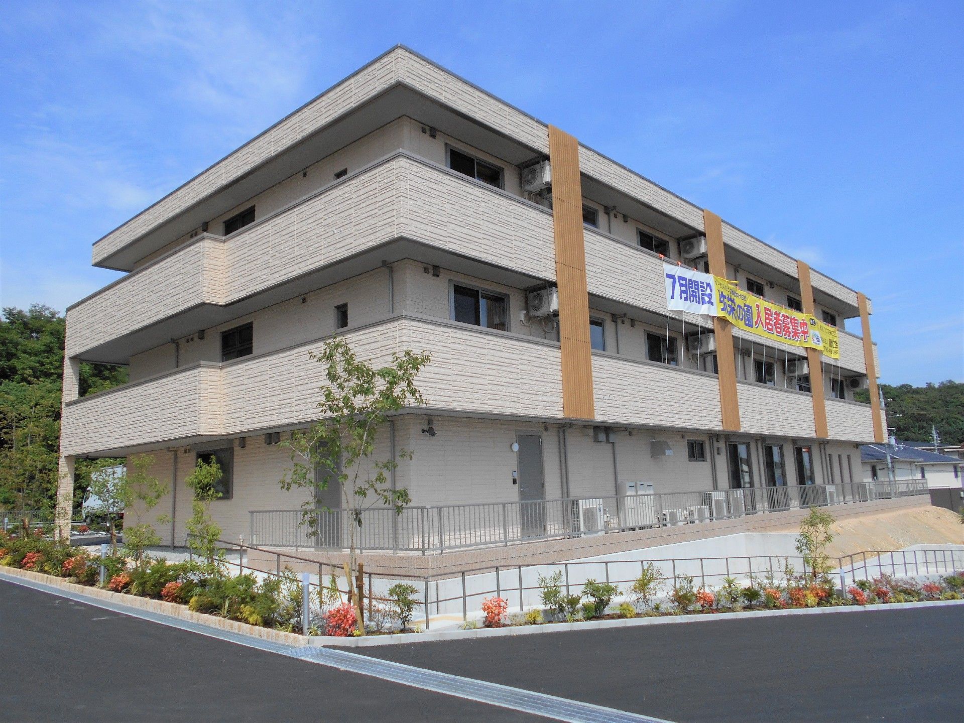 ３階建てのグレー系の建物。屋上には柵が付き、建物周辺には花や木が植えられている。