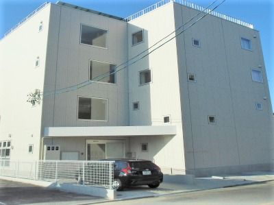 ホワイト基調の鉄骨造の建物の写真。マンション風の外観で雨除けと駐車場スペースがあるのがわかる