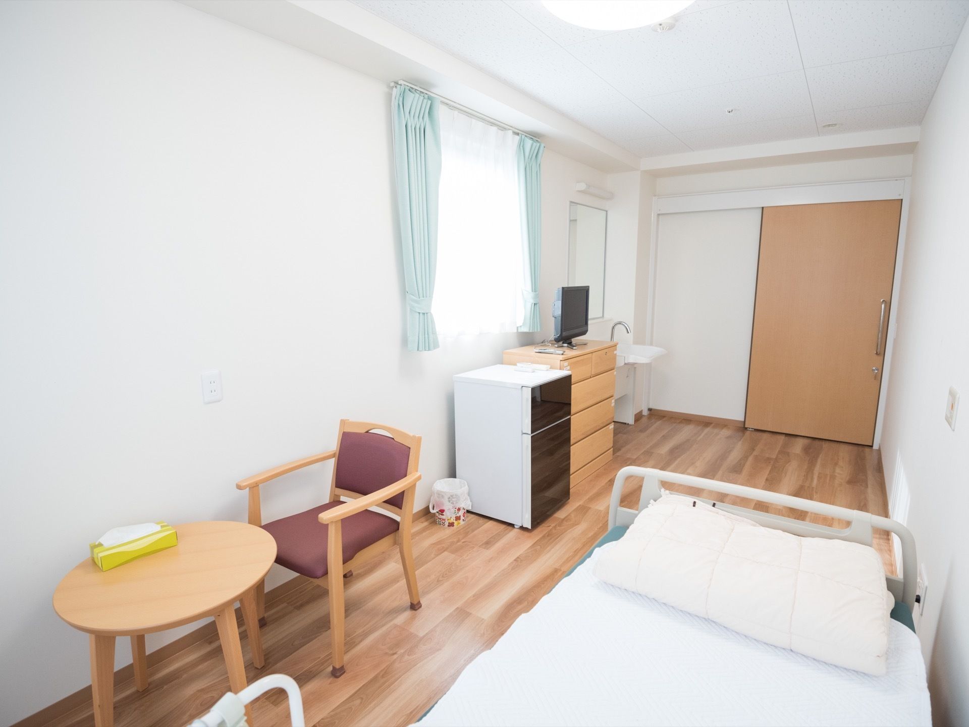 大きな引き戸の居室である。ベッドが１台置かれ、窓の下には、木製のチェストと冷蔵庫が並べてある。その横には、ゴミ箱が置いてある。