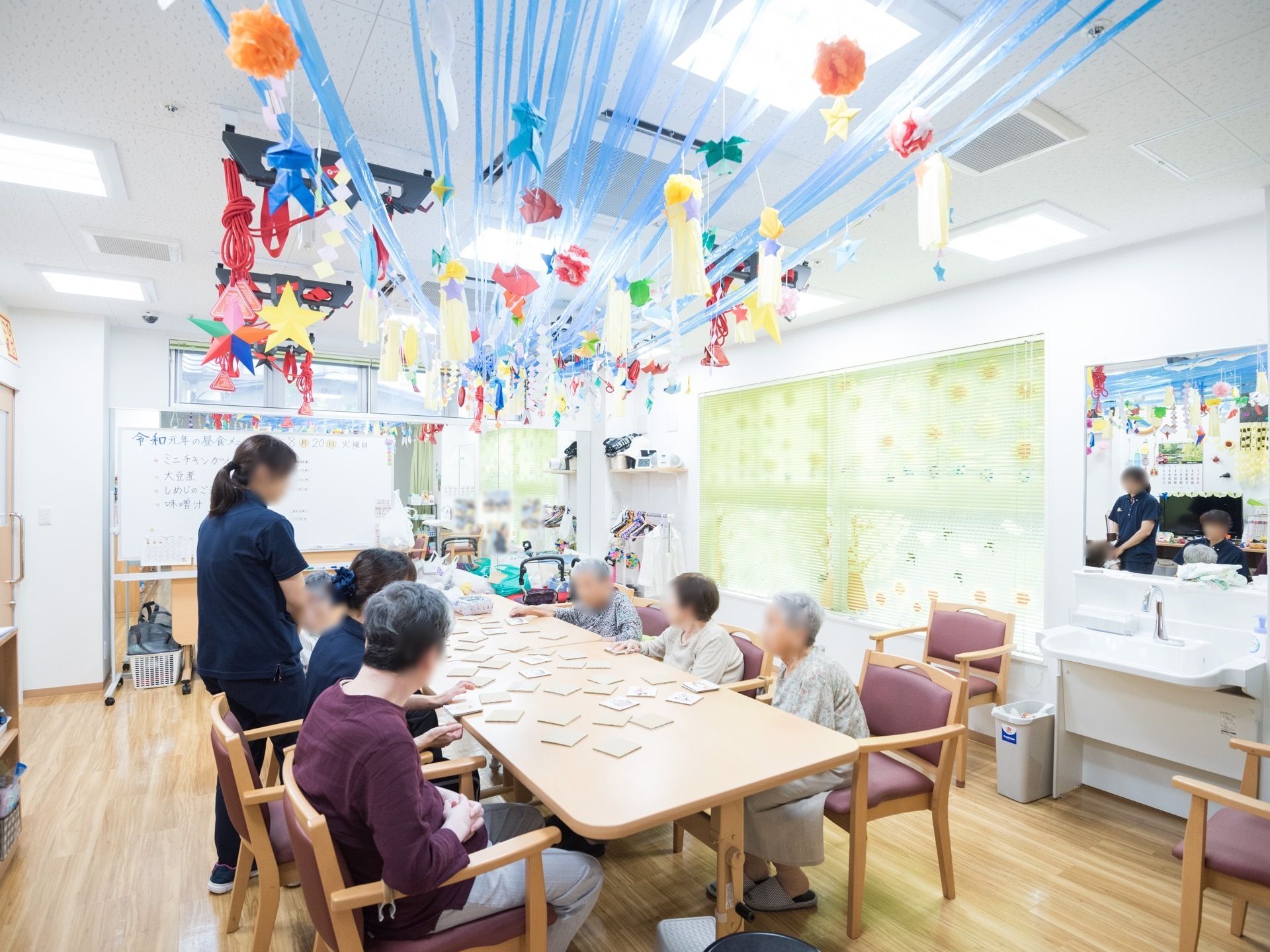 中央に置かれた大きなテーブルを囲むように、複数の入居者様が座っているようすである。テーブルの上にはたくさんの正方形のものが置かれている。