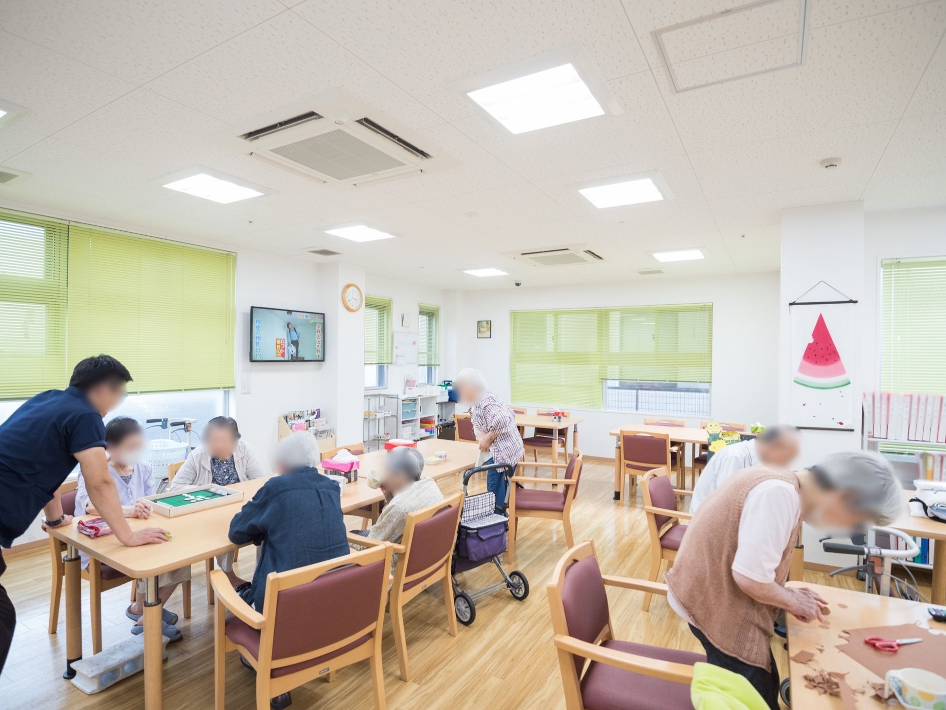 並べられたテーブルで、入居者様が制作や、ゲームをしている様子である。窓には、緑色のブラインドがかかっている。