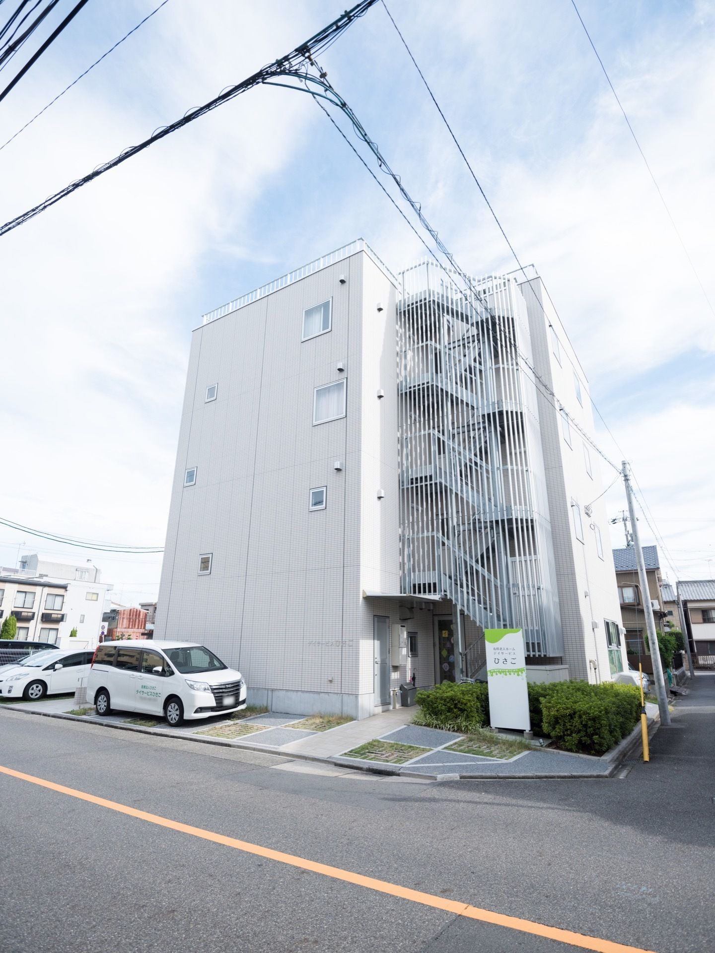 コンクリート造りの４階建ての建物である。外には、螺旋状の非常階段が設置されており、建物横見は車が１台停まっている。