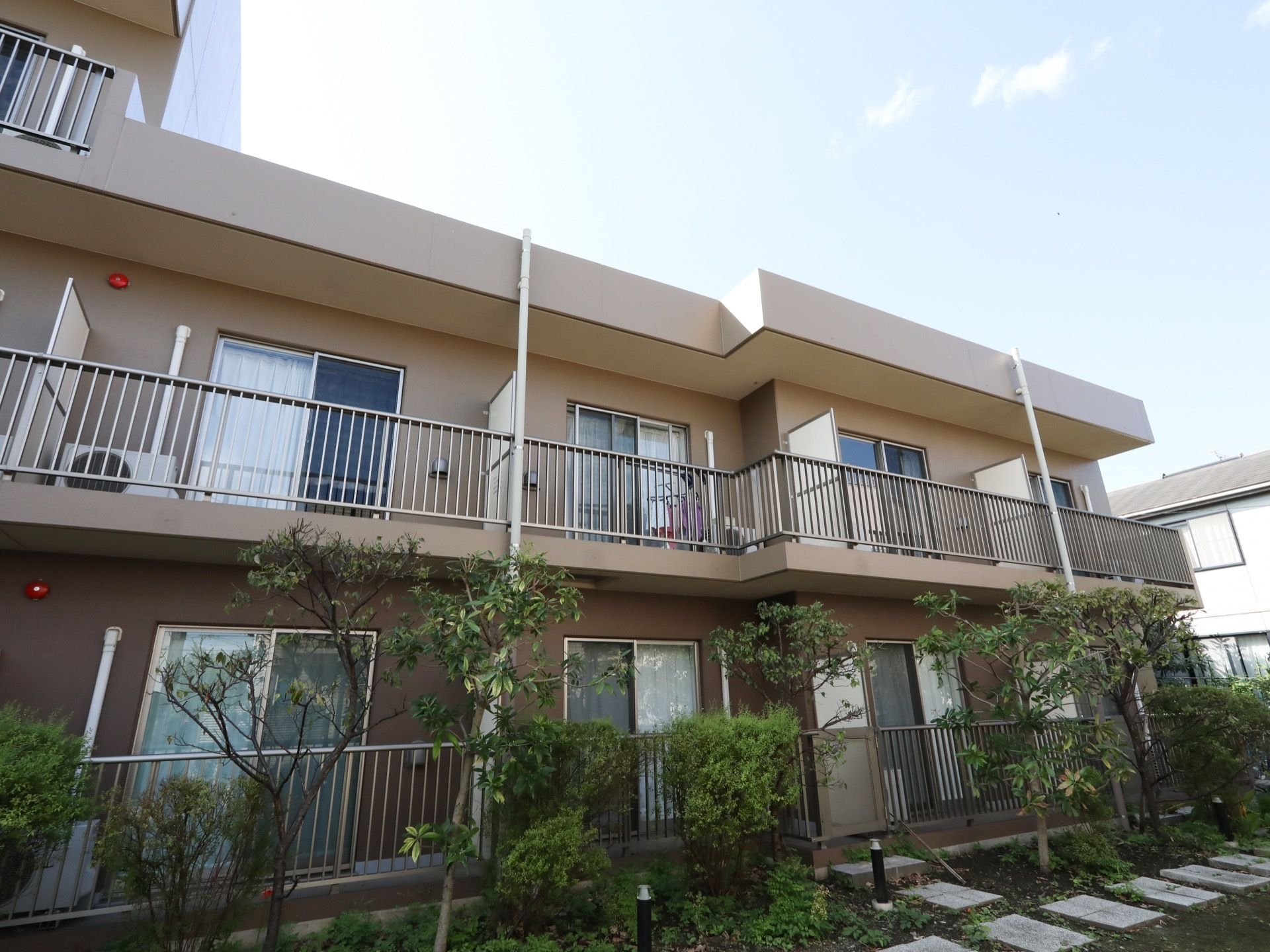住宅地の一角にマンションタイプの建物が建っている。敷地は植栽に囲まれていて、居室にはベランダが付いている。
